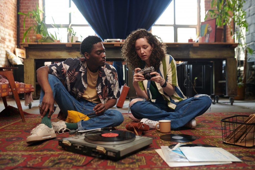 two young people sitting on the floor listening to a record on a record player and looking at an analog camera